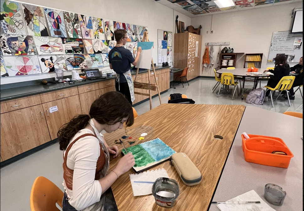 two students painting in art class — one is seated at a table, and another is standing nearby at an easel. They are surrounded by art on the walls.