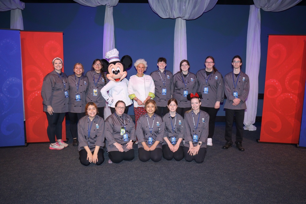 A group of culinary arts students pose in gray chef's jackets with Mickey Mouse and chef Anne Burrell standing in the middle of the group