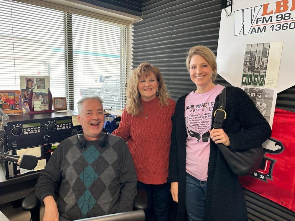 The host of WLBK radio is seated next to a smiling Christi Volkening and Tara Wilkins of Genoa-Kingston schools inside the radio studio.